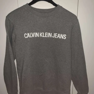 Grå sweatshirt från Calvin Klein Jeans - Säljer en stilren grå sweatshirt från Calvin Klein Jeans med vit logotyp på bröstet. Tröjan har långa ärmar och en rund halsringning. Perfekt för en avslappnad look. Står xs men sitter som S/M