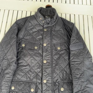 Grå quiltad jacka från Barbour - Säljer en stilren grå quiltad jacka från Barbour. Jackan har en hög krage med knappar och flera praktiska fickor framtill. Perfekt för kyligare dagar.