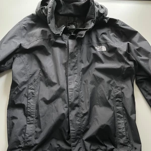 Svart vindjacka från The North Face - Säljer en svart vindjacka från The North Face. Jackan har en huva. Perfekt för blåsiga dagar och lätt att packa med sig. The North Face-loggan syns tydligt på bröstet och ryggen. Små skönhetsfel finns på jackan men inget man tänker på. 