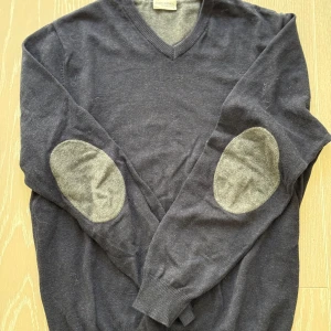 Mörkgrå tröja från John Henric - Säljer en stilren mörkgrå tröja från John Henric med v-ringning och kontrasterande ljusgrå armbågslappar. Perfekt för en elegant och avslappnad look. Tröjan har långa ärmar och ribbade muddar.