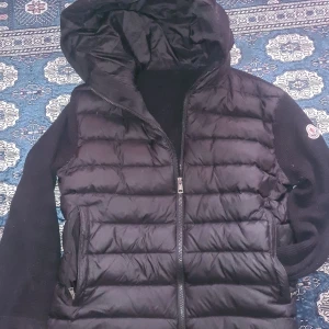 Svart moncler cardigan  - Säljer en snygg svart cardigan från Moncler med huva och dragkedja. Cardigen har ett stilrent quiltat mönster och är perfekt för kyligare dagar. Märkesloggan syns tydligt på sidan. Passar bra för både vardag och mer avslappnade tillfällen.