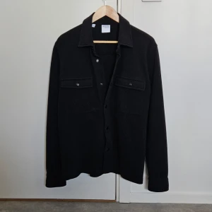 Svart overshirt från Selected Homme - Snygg svart overshirt från Selected Homme med knappar framtill och två bröstfickor med lock. Perfekt för lager-på-lager-stil. Tillverkad i ett mjukt material som ger en bekväm passform. Använd ett fåtal gånger så därför väldigt bra skick!