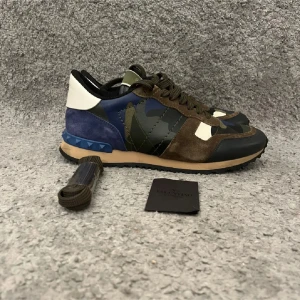 Valentino Rockrunner i camouflage - Snygga Valentino Rockrunner sneakers i ett coolt camouflage-mönster. Skorna har en mix av material som mocka och skinn i färgerna grön, blå, vit och brun. De har en unik design med blå nitar på hälen och en bekväm gummisula. Storlek 40,5 passar 41,5 