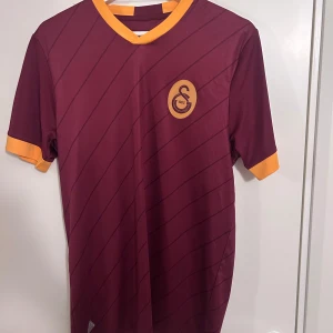 Galatasaray träningströja - Snygg vinröd fotbollströja från Adidas med diagonala ränder och gulddetaljer vid krage och ärmslut. Perfekt för fotbollsfans som vill sticka ut på planen eller läktaren. Tröjan har korta ärmar och en stilren design med klubbemblem på bröstet.