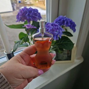 Jean Paul Gaultier 50 ml - Elegant parfym i en flaska formad som en kvinnokropp. Flaskan är genomskinlig med en varm, bärnstensfärgad vätska inuti. Den har en silverfärgad spraypump. Perfekt för den som vill ha en unik och stilfull doftupplevelse. Bara prov använd.