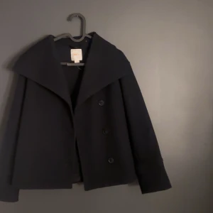 Mörkblå kappa från Gina Tricot - Elegant marinblå kappa från Gina Tricot med stora knappar framtill. Den har en bred krage och långa ärmar, perfekt för kyligare dagar. En stilren och tidlös design som passar till många tillfällen.