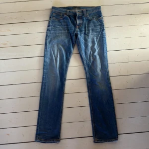 Blå jeans från Nudie Jeans - Säljer ett par klassiska blå jeans från Nudie Jeans. De har en slim fit passform och är tillverkade i slitstarkt denim. Perfekta för en avslappnad stil med snygga detaljer som orange sömmar och en läderpatch med logga på baksidan.