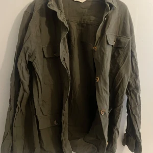 Olivgrön jacka med knappar - Säljer en olivgrön jacka med knappar framtill och två bröstfickor. Jackan har en avslappnad stil och är perfekt för lager-på-lager. Den är långärmad och har en klassisk krage. Klippt nertill