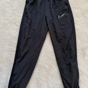 Svarta träningsbyxor från Nike - Säljer ett par svarta träningsbyxor från Nike med Dri-FIT-teknologi. Byxorna har en normal passform och är utrustade med dragkedjor vid fickorna. Perfekta för träning och andra sportaktiviteter. Storlek M
