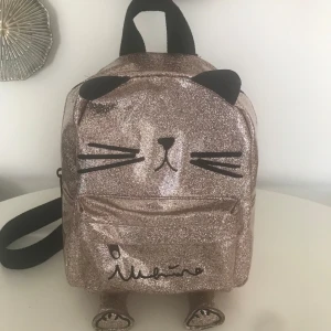 Glittrig ryggsäck med kattmotiv - Säljer en superfin glittrig ryggsäck i guld med ett gulligt kattmotiv. Väskan har svarta detaljer som öron och morrhår, samt justerbara axelband. Perfekt för att ge lite extra bling till din outfit!