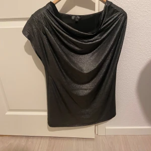 Svart glittrig topp från KappAhl - Snygg svart glittrig topp från KappAhl med draperad halsringning. Perfekt för en kväll ute eller festliga tillfällen. Toppen har en elegant och stilren design.