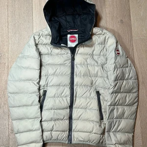 Beige dunjacka från Colmar - Snygg beige dunjacka från Colmar med huva och dragkedja. Jackan har en quiltad design och två praktiska fickor med dragkedja. Perfekt för kyligare dagar.