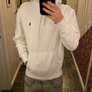Vit hoodie från Ralph Lauren - Säljer en stilren vit hoodie från  Ralph Lauren med det klassiska logotypbroderiet på bröstet. Tröjan har en känguruficka och justerbar huva med snören. Perfekt för en avslappnad look.