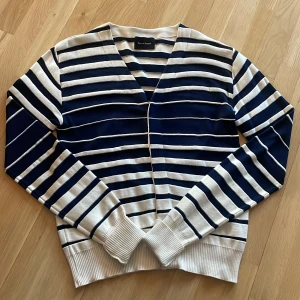 Randig kofta från Nom de Guerre - Exklusiv randig kofta från Nom de Guerre i mörkblå samt vit färg. Tillverkad i en mjuk blandning av bomull och silke. Koftan har en v-ringad design och långa ärmar, perfekt för en klassisk look. Nypris: 2 500kr