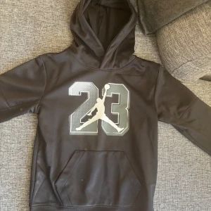 Svart hoodie från Jordan - Säljer en svart hoodie från Jordan med det ikoniska Jumpman-logotypen och siffran 23 på framsidan. Tröjan har en stor ficka framtill och en bekväm huva. Perfekt för sportiga tillfällen eller en avslappnad stil.