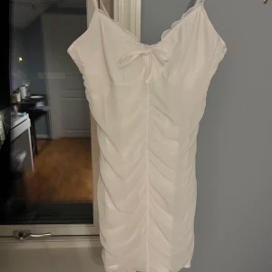 Vit klänning med rosett - Säljer en stilren vit klänning med smala axelband och en söt rosett framtill. Klänningen har en åtsittande passform och rynkade detaljer som ger en fin struktur. Perfekt för sommarens alla tillfällen! Är köpt på H&M och jag TROR det är storlek M men är ej helt säker då lappen inte är kvar. Skriv gärna om ni undrar nått!