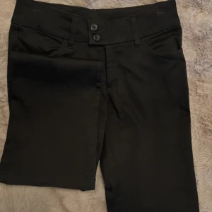 Svarta lågmidjade kostymbyxor med knappar - Snygga svarta lågmidjade kostymbyxor med två knappar och dragkedja framtill. Byxorna har en klassisk design med hällor för bälte och raka ben. Perfekta för en stilren look.