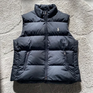 Svart dunväst från Ralph Lauren Sport - Snygg svart dunväst från Ralph Lauren Sport med dragkedja och broderad logga på bröstet. Västen är quiltad och perfekt för kyligare dagar. Tillverkad i polyester med dunfyllning för extra värme. 