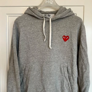 Grå hoodie från Comme des Garçons - Säljer en stilren grå hoodie från Comme des Garçons med det ikoniska röda hjärtat på bröstet. Storlek är M men den är lite mindre i storleken så skulle säga att den sitter som S