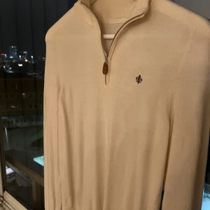 Morris/Beige tröja med dragkedja - Säljer en snygg beige tröja med halv dragkedja och broderad detalj på bröstet. Perfekt för en stilren look. Tröjan har långa ärmar och en hög krage. Nytt pris ca 1300 kr, Mitt pris endast 400 kr!