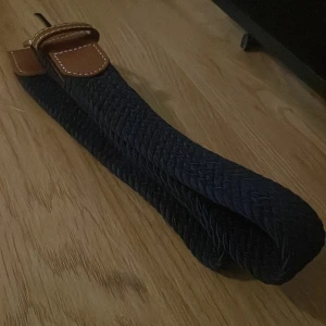 Mörkblått flätat bälte  - Snyggt flätat bälte i mörkblått med bruna skinnliknande detaljer och en silverfärgad spänne. Perfekt för att ge en stilren touch till din outfit.