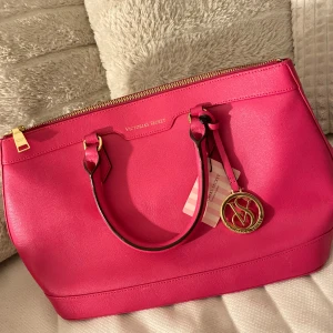 Rosa handväska från Victoria's Secret - Snygg rosa handväska från Victoria's Secret med gulddetaljer och en praktisk dragkedja upptill. Väskan har två handtag och en dekorativ VS-logga i guld. Perfekt för att ge din outfit en färgklick! ÄR I ÄKTA LÄDER!!