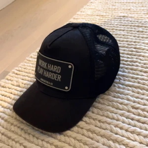 John hatter keps  - Snygg svart keps med texten 'Work Hard Play Harder' framtill. Kepsen har nätpaneler på sidorna och baksidan för extra ventilation. Perfekt för en avslappnad stil. 