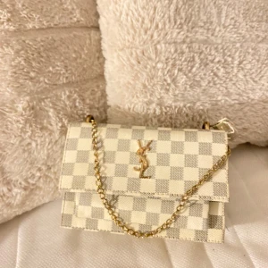 Rutig handväska  - Snygg rutig handväska med kedjerem och ikonisk logotyp på framsidan. Väskan har en elegant design med en kombination av beige och vit färg. Perfekt för att ge en lyxig touch till din outfit.