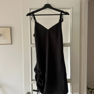 Svart axelbandslös klänning - Snygg svart oanvänd klänning från NAKD med justerbara axelband och en  drapering på sidan. Slutar ovanför knäna! 