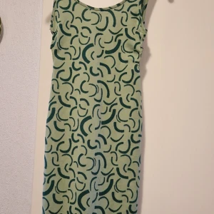 Grön mönstrad klänning från Bershka - Säljer en snygg grön klänning från Bershka med ett unikt mönster i mörkare nyanser. Klänningen är ärmlös och har en rund halsringning. Perfekt för en stilren look!