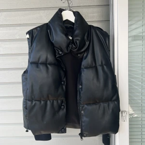 Svart dunväst från Zara - Snygg svart dunväst från Zara med en stilren design. Västen har en hög krage och stängs med både dragkedja och knappar. Perfekt för att hålla värmen under kyliga dagar.