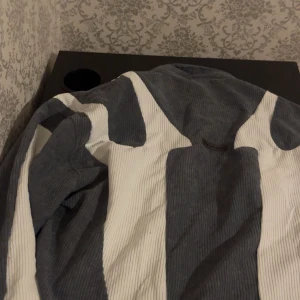 Grå och vit jacka med dragkedja - Snygg grå och vit jacka med dragkedja framtill. Jackan har ett unikt mönster och är tillverkad i ett ribbat material som ger en cool look. Perfekt för att sticka ut i mängden!