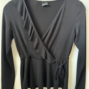 Svart bus/tröja från Gina Tricot - Snygg svart tröja från Gina Tricot med volangdetaljer och knytning i midjan. Toppen har långa ärmar och en v-ringad design som ger en elegant look. Perfekt för att klä upp eller ner beroende på tillfälle.