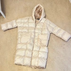 Beige pufferjacka från WPR - Säljer en beige pufferjacka från WPR. Jackan har en lång design med huva och stora fickor framtill. Perfekt för kyliga dagar med sin quiltade stil och långa ärmar. En stilren och praktisk jacka för vintern.