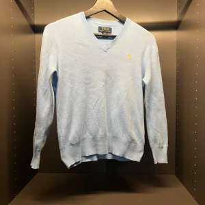 Ljusblå v-ringad tröja från Polo Ralph Lauren - Säljer en stilren ljusblå v-ringad tröja från Polo Ralph Lauren. Tröjan har en broderad gul logotyp på bröstet och är tillverkad i ett mjukt material. Perfekt för en avslappnad men elegant look.