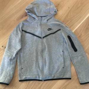 Grå huvtröja från Nike - Säljer en grå barn Nike tech fleece med dragkedja och huva. Tröjan har svarta detaljer och en ficka med dragkedja på ärmen. Perfekt för en sportig look eller avslappnade dagar.  storleken är 147-158 men är passar en storlek upp.