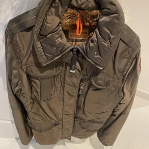 Brun jacka från Parajumpers - Säljer en brun jacka från Parajumpers med pälsfoder och flera fickor. Jackan har en dragkedja och en broderad patch på ärmen. Perfekt för vintern med sin varma och skyddande design.