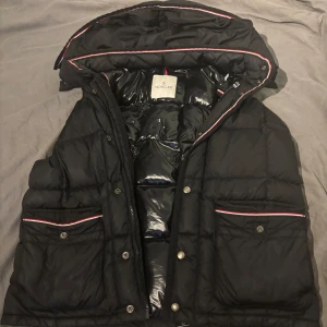 Svart dunväst från Moncler - Strl 4.   Säljer en snygg svart dunjacka från Moncler med avtagbar huva och dragkedja. Jackan har långa ärmar och praktiska fickor med knappar. Den har en stilren design med röda och vita detaljer längs kanterna.