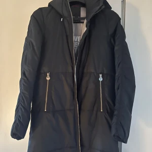 Svart dunjacka från Röhnisch - Snygg svart lång dunjacka från Röhnisch med dragkedja och två fickor med dragkedjor framtill. Jackan har en hög krage och långa ärmar, perfekt för kyliga dagar. Den är stilren och funktionell, idealisk för både stad och natur.