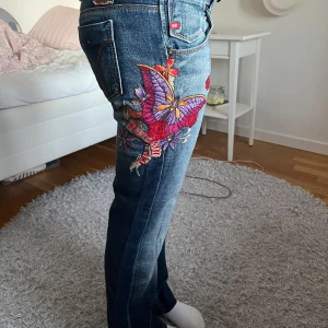 Broderade lågmidjade miss sixty jeans - Skit coola lågmidjade Miss Sixty jeans med broderade fjärilar och blommor i lila och rött på sidan. De har en blå tvätt och en lätt bootcut. Dem är strlk 27 och midjemåttet är 39 och innerbenslängden 72. Säljer för att de är försmå för mig! 🥰