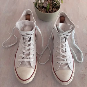 Vita Converse Chuck Taylor All Star - Klassiska vita Converse Chuck Taylor All Star med snörning och rund tå. Skorna har den ikoniska logotypen på sidan och en röd rand längs sulan. Perfekta för en avslappnad stil.