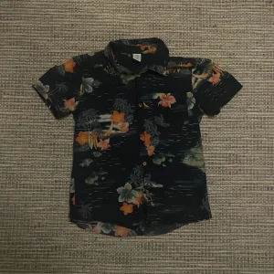 Mönstrad kortärmad skjorta - Säljer en cool kortärmad skjorta med tropiskt mönster i orange, grön och svart. Perfekt för sommarens äventyr. Skjortan har knappar framtill och en bröstficka.          Original priset är 300