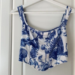 Vit och blå croppad linnetopp - Säljer en croppad linnetopp med breda axelband och ett unikt blått botaniskt mönster på vit bakgrund. Toppen har en lätt böjd nederkant. Materialet är mjukt och svalt, perfekt för varma dagar. Aldrig använd. Väldigt stretchig 