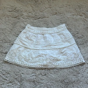 Vit kortkjol med broderade detaljer - Supersöt vit kortkjol med broderade blommor och spetsdetaljer. Kjolen har en smockad midja som gör den bekväm och lätt att bära. Perfekt för varma dagar och ger en fräsch och somrig vibe till din outfit. Storlek 9-10 men passar 158,164 