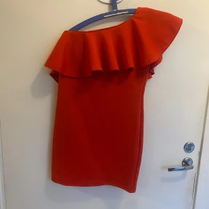 Röd kortklänning med volang - Säljer en snygg röd kortklänning med en stor volang upptill och oneshoulder-design. Klänningen har en clean look utan mönster och är perfekt för dig som gillar att sticka ut. Materialet känns mjukt och har lite struktur, vilket gör den bekväm att bära. Passar dig som vill ha något trendigt och annorlunda.