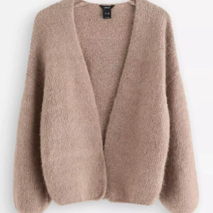 Beige stickad kofta från Lindex - Mysig och loose stickad kofta från Lindex i en mjuk beige ton. Koftan har långärmade, lite puffiga ärmar och en öppen front utan knappar. Perfekt att slänga över en t-shirt för en chill look. Materialet är fluffigt och känns lyxigt mot huden.