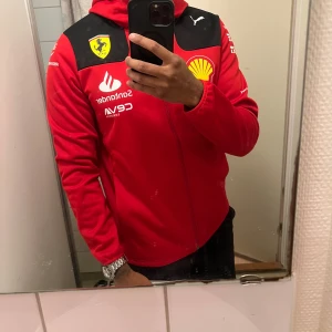 Ferrari F1 Puma röd hooded jacka M - Säljer en röd Ferrari F1 Puma hooded jacka med svarta detaljer och tryck från Shell, Santander, Pirelli och fler sponsorer. Jackan har hel dragkedja, huva och är långärmad. Materialet är mjuk polyester, perfekt för dig som gillar racing och sportig stil. Skriv vid intresse!