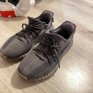 Adidas Yeezy Boost 350 V2 Cinder - Säljer ett par Adidas Yeezy Boost 350 V2 i mörkgrå färg. Skorna har stickad ovandel i syntet, snörning och en ribbad platt sula med Boost-dämpning. Ikoniska tre ränder på hälen och cool textur på ovandelen. Perfekt för dig som gillar streetwear och vill sticka ut.