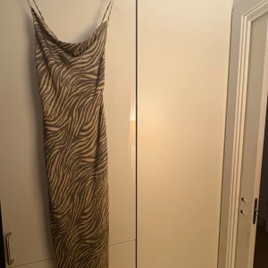 Adoore klänning  - Säljer en snygg långklänning med zebramönster i beige och brunt. Klänningen har tunna axelband, öppen rygg med snörning och en smickrande siluett. Perfekt för dig som vill sticka ut med ett djuriskt print och coola detaljer.