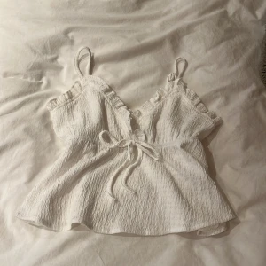 Vit linnetopp med volang och knyt - Supersöt vit linnetopp med smala axelband, volangkant vid v-ringningen och knyt i midjan. Toppen har en lätt rynkad struktur och ärmlös design, perfekt för varma dagar. Luftig och flowy passform som ger en chill vibe.